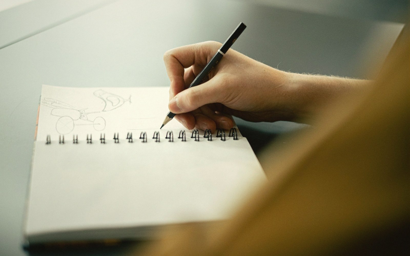 A hand sketches a vehicle design with pencil in a spiral-bound notebook
