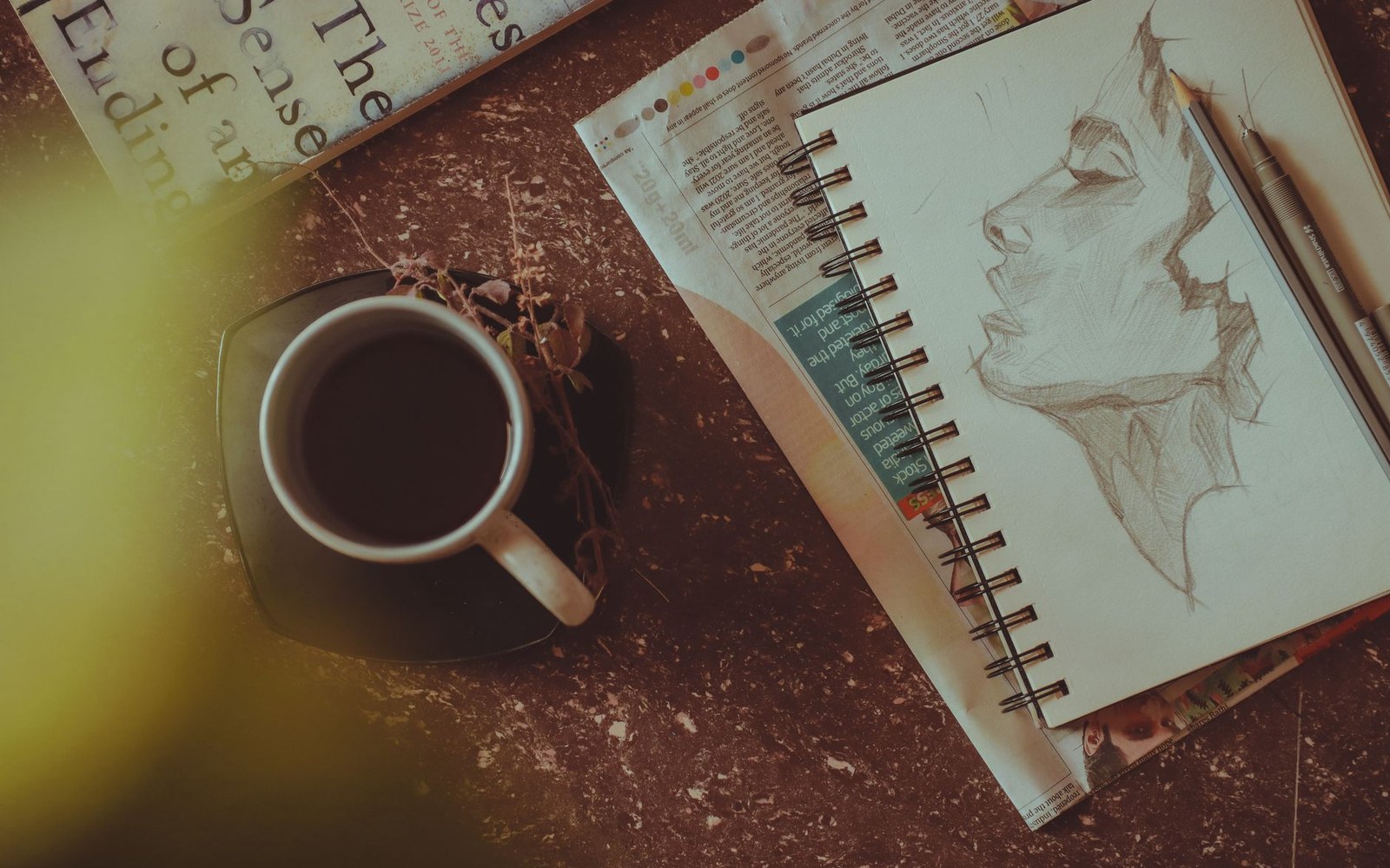 A pencil sketch of a face in a spiral notebook beside a coffee cup and book