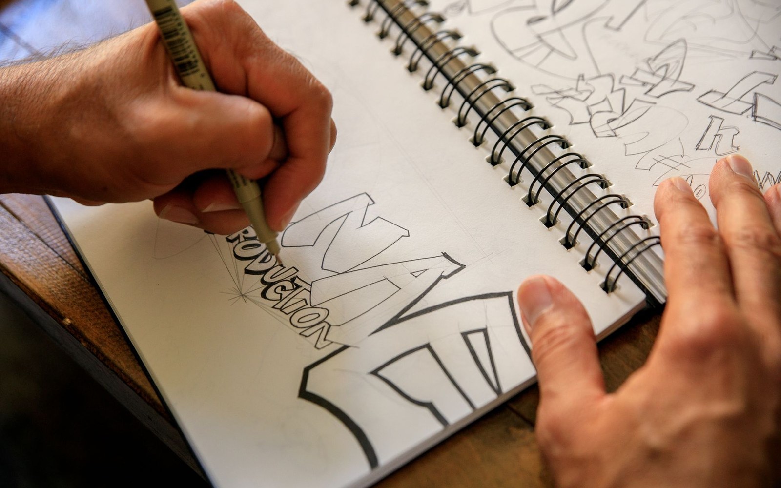 Hands sketching bold graffiti-style block lettering in a spiral-bound notebook