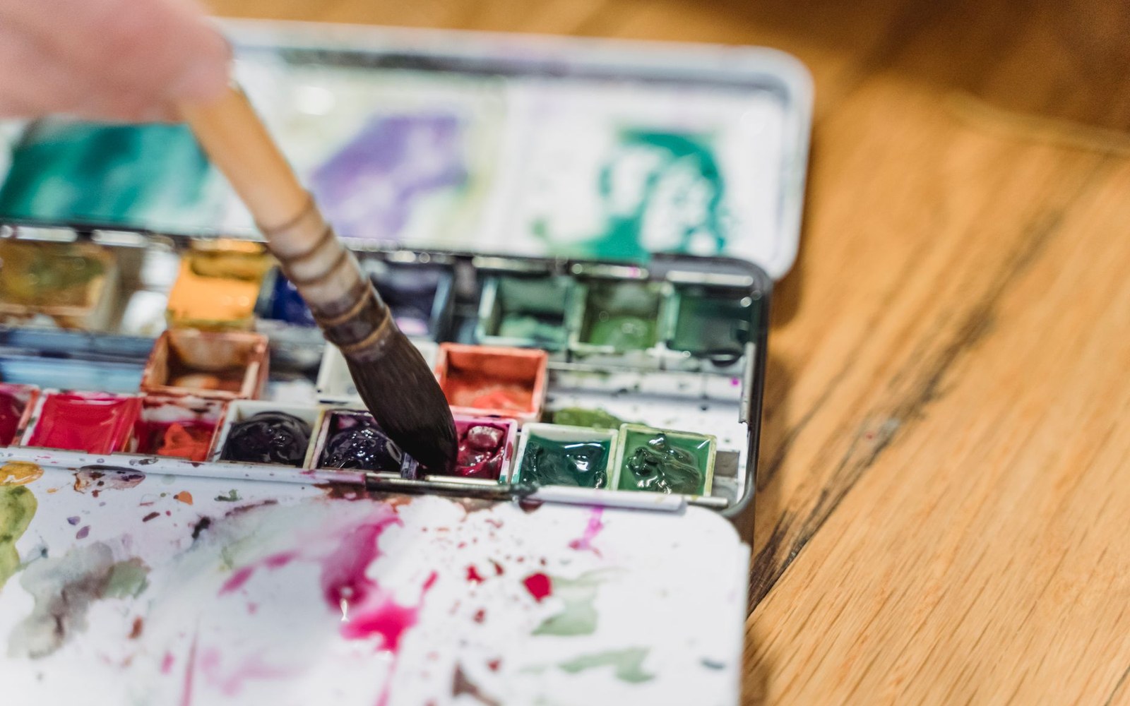A brush dips into a tin watercolor palette with rows of dried paint pans