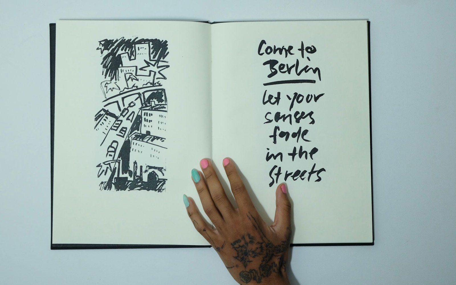 An open sketchbook with a city skyline drawing and handwritten text reading "come to Berlin, let your senses fade in the streets," a tattooed hand resting on the page
