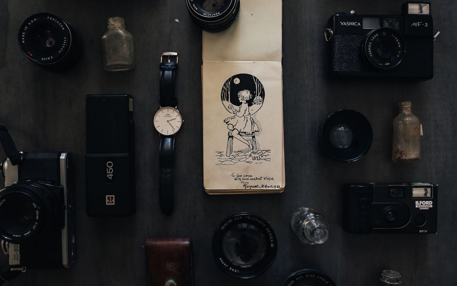 Vintage cameras, lenses, a wristwatch and glass bottles arranged around an illustrated notebook